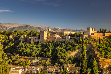 Fototapeta premium Panorama of Alhambra from the Mirador de San Nicolas viewpoint in Granada town in Spain at sunset. Ancient palace nestled in a lush green hill, overlooking a town and mountain range under a blue sky.