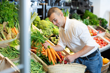 Attentive young man purchaser holding organic carrots at the counter in large supermarket