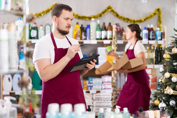 Focused bearded man seller restock of goods using tablet, female assistant laying out goods in background in small store decorated for New Year holiday