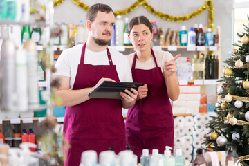 Bearded man and woman sellers restock of goods using tablet in small store decorated for New Year holiday