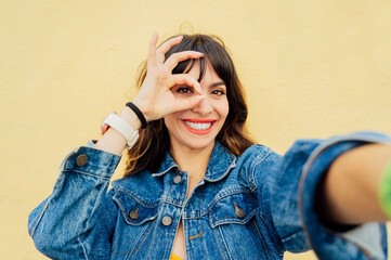 Young woman making ok sign taking selfie with yellow background