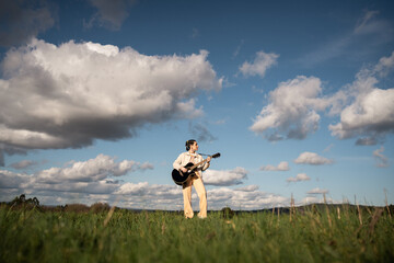 Obraz premium Woman playing acoustic guitar in grassy field beneath blue sky, white clouds surrounding her peaceful musical moment in sunlit outdoor setting