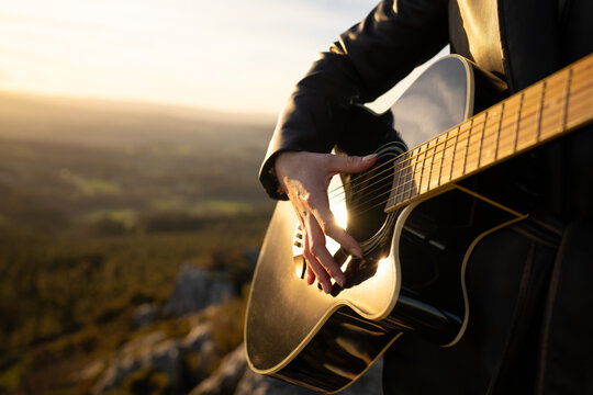 Female guitarist strumming acoustic instrument at golden hour, silhouetted against dramatic wilderness backdrop, embodying musical inspiration and creative energy