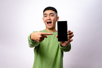 An Asian man in a green sweatshirt is smiling and pointing at a smartphone with a blank screen. He...