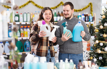 Cheerful married couple customer buying spray and detergent in small store decorated for New Year holiday