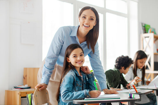 Happy teacher working in private school, standing near school girl and both smiling at camera in classroom interior, free space