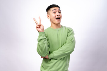 An Asian man in a green sweatshirt is making a peace sign with his right hand while slightly smirking. His arms are crossed, and he stands against a white background, conveying a confident