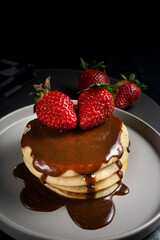 A stack of pancakes with chocolate sauce and strawberries on a gray plate, placed on a black leather mat 