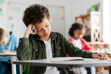 Upset tired Latin boy student of elementary school trying to solve mathematical problems or writing test in classroom