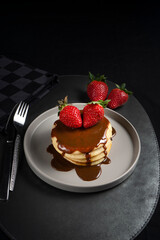 A stack of pancakes with chocolate sauce and strawberries on a gray plate, placed on a black leather mat with a fork and knife beside a checkered napkin.