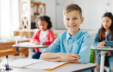 Portrait of positive schoolboy sitting at school desk looking and smiling at camera during lesson, classmates on background