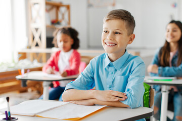 Happy smart schoolchildren listening carefully to teacher at the lesson, sitting at desks in classroom, focus on smiling boy. New academic semester year or end of educational year