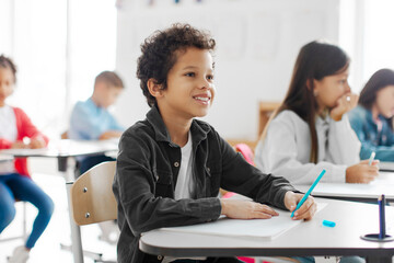 Smart Latin boy listening teacher and writing in notebook, taking notes or writing final test in private school classroom
