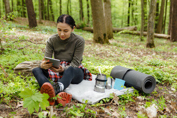 Young woman hiker sitting on a blanket in a serene forest, using a tablet next to her camping gear, blending the joys of nature with the convenience of technology
