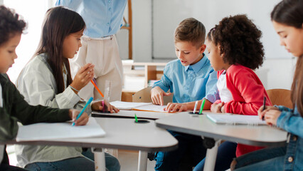 Group of preteen children studying with teacher in classroom at elementary school, having group...