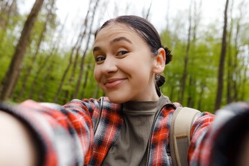 Happy young female hiker taking a selfie while enjoying a vibrant walk through a lush green forest,...