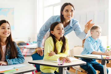 Positive female teacher pointing at blackboard and explaining while standing among pupils, creating an interactive learning atmosphere