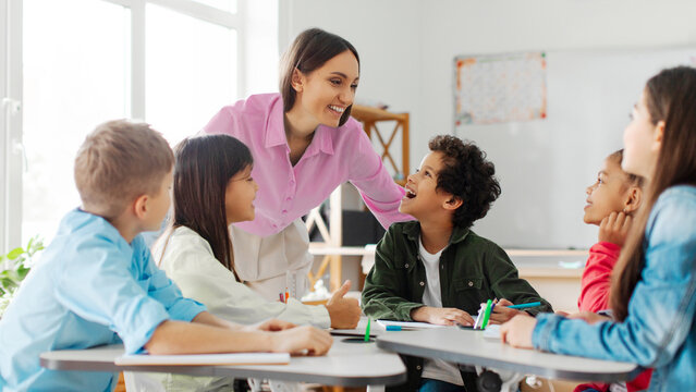 Cheerful teacher talking to kids, children sitting at desks in classroom and listening their teacher with interest, panorama - Powered by Adobe