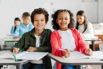 Happy black friends schoolboy and schoolgirl smiling at camera, sitting at desks in classroom with other children on background
