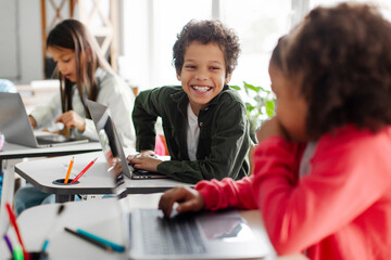 Latin boy chatting with girl during lesson, sitting in school classroom with laptops on desk. Classmates study in classroom. Modern technology education