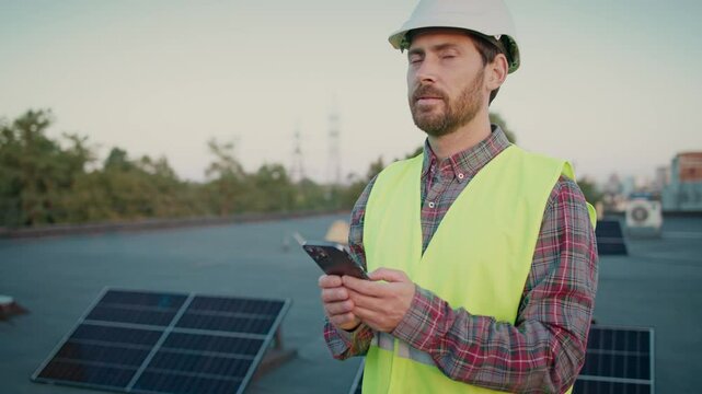 Male engineer on rooftop tapping on mobile phone at solar panels. Combining technology with sustainable energy. A modern approach to renewable innovation. Man texting and scrolling on smartphone.
