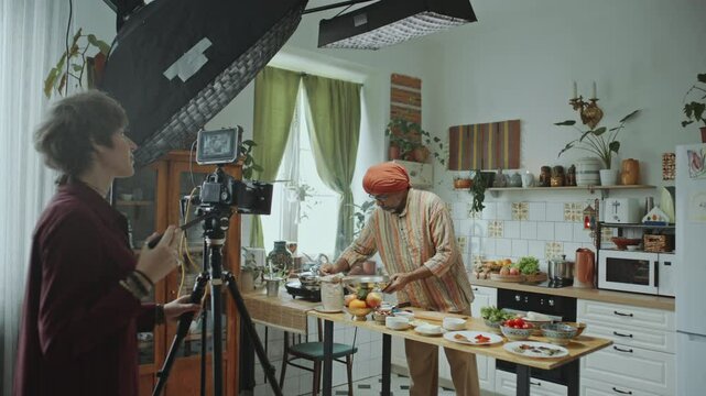 Female filmmaker recording senior Indian chef in turban preparing meal in kitchen with professional camera and lighting setup, creating video content for culinary vlog