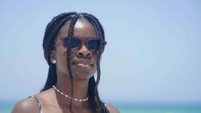 Portrait of cheerful woman with braided hair and sunglasses smiles brightly during a sunny day at the beach, slow motion