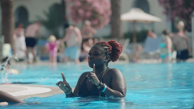 Cheerful young black woman dances playfully in a sunny pool area, surrounded by people enjoying leisure and summer vibes