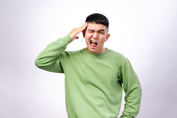 An Asian man wearing a green sweatshirt against a white background is clutching his head with both...