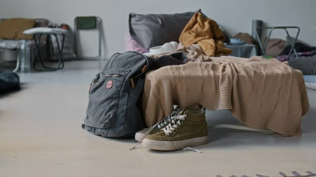 No people medium closeup of backpack, pair of sneakers on floor, next to folding bed with pillow and blankets, in refugee shelter for homeless people displaced due to war or natural disaster