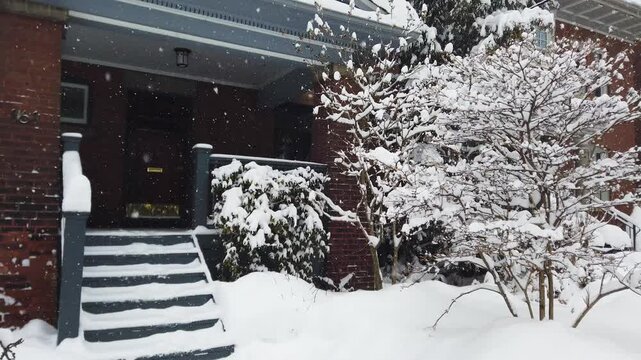 Snowy winter in Toronto, Ontario, Canada. A beautiful small yard with bushes covered with thick snow near a brick house with a porch during a snowfall.