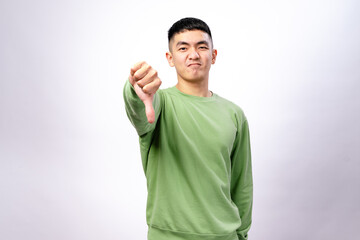 An Asian man in a green sweatshirt stands against a white background, making a thumbs-down gesture with a displeased facial expression, expressing disapproval or dissatisfaction