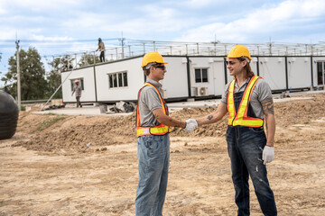 Two construction workers wearing hard hats and safety vests shaking hands at a construction site,...