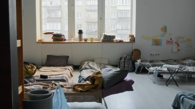 Full dolly shot of bedroom in refugee shelter or camp, for people displaced by war, violence or natural disaster, with mattresses on floor, stacks of blankets, bags, childs drawings on wall at daytime