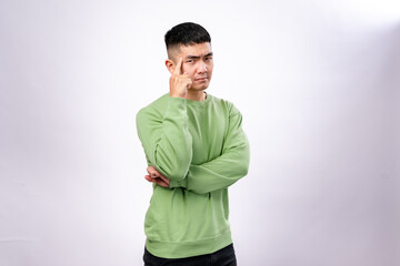 An Asian man in a green sweatshirt poses thoughtfully with a finger on his temple and a serious...