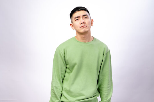 An Asian man in a green sweatshirt stands against a white background, slouching with a tired, exhausted, or disappointed expression, his face showing fatigue, frustration, or sadness