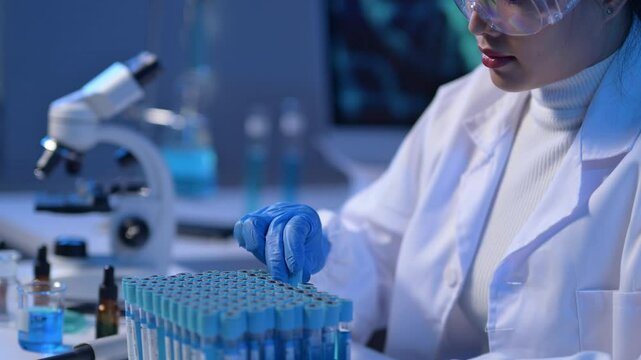 A scientist is working with a tray of test tubes. The tubes are blue and white and are arranged in rows. The scientist is wearing gloves and a lab coat. Scene is serious and focused