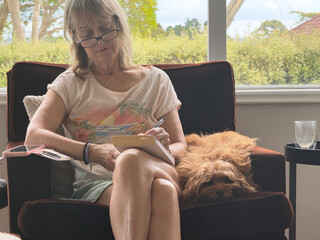 A lady sits in shorts beside her pet dog as she writes in a book.A window behind has a view of greenery and trees.