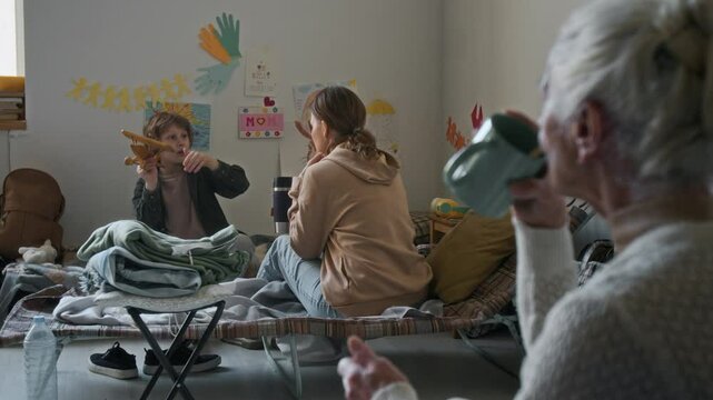 Full shot of cheerful Caucasian boy sitting on folding bed in emergency accommodation for natural disaster victims, playing with toy plane, chatting to mom, elderly lady drinking tea in foreground