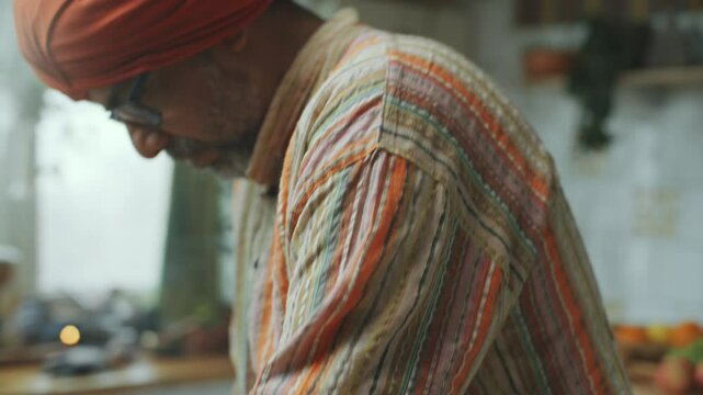 Senior Indian man in kurta and turban adding water while kneading dough in metal bowl on wooden kitchen table with various cooking ingredients on it. Close-up shot