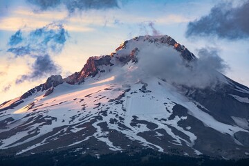 Majestic Mount Hood at sunrise, snow-capped peak shrouded in clouds. Sunrise hues paint the...