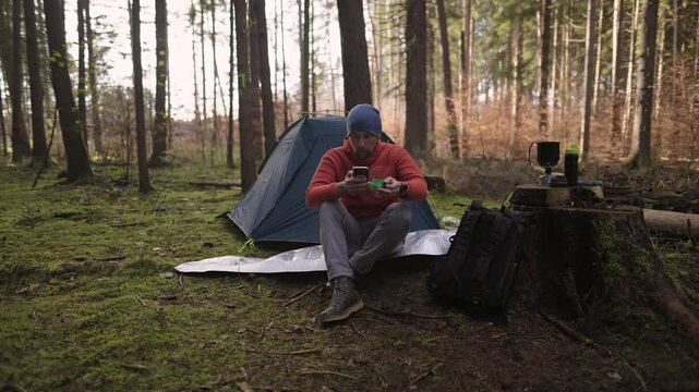 Male hiker drinking tea and surfing Internet on smartphone in tent camp during hiking in fall in woods. Backpacker makes route in map in phone and drinks hot drink from cup in background of tent. 