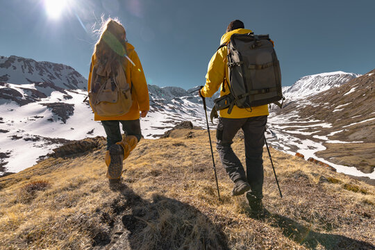 Two hikers with backpacks on their backs are walking along a trail with a view of snowy mountains and blue sky. A walk in the mountains, a view from the back. - Powered by Adobe