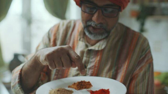Indian culinary blogger in turban pointing at assorted ground spices on white plate, explaining how to use them in cooking as filming food vlog in kitchen