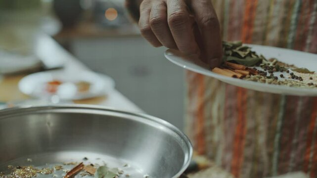 Hands of male chef adding whole spices like bay leaves, cinnamon sticks and peppercorns to heated pan for cooking Indian food in kitchen. Close-up shot