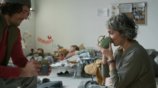 Medium shot of young Caucasian man in warm vest and beanie sharing cup of hot tea with mature woman, thanking him, while staying together in refugee camp during natural disaster