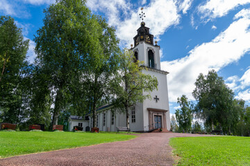 Naklejka premium A tranquil day at the Church of Sweden in Idre Dalarna, showcasing the building’s architecture amidst green trees under a blue sky and white clouds. Visitors enjoy the peaceful atmosphere