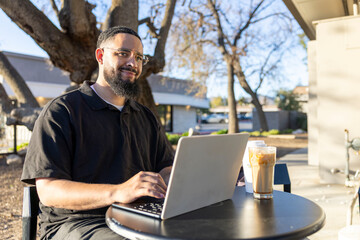 Fototapeta premium A man is sitting outside at a small table, deeply engrossed in his laptop while also enjoying a cup of coffee, finding inspiration from the lush greenery surrounding him on this gorgeous, sunny day