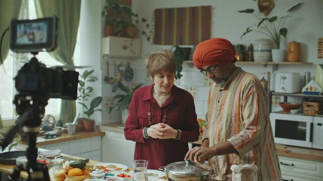 Female food blogger and Indian chef in turban cooking traditional dish and commenting on ingredients in kitchen with camera setup, filming culinary vlog together