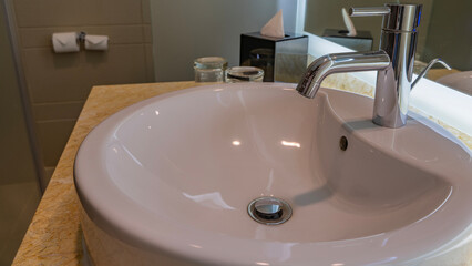 Rounded white ceramic washbasin. The drain hole is in the center. A shiny metal faucet on the edge. There are glasses and a box of napkins on the marble countertop. Reflection in the wall mirror. 
